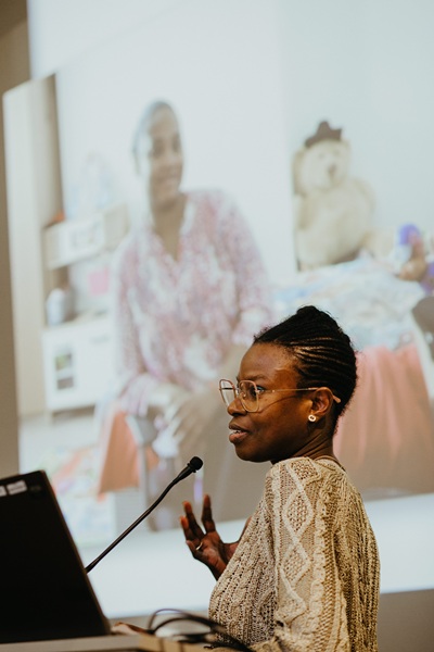 Woman speaking from a podium, gesturing towards a photo of a seated woman, projected on the screen behind her.