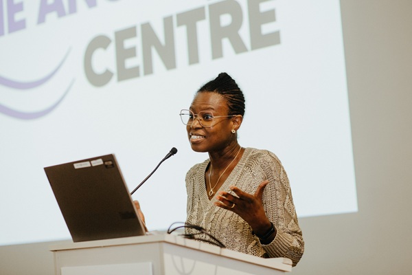 Woman speaking from a podium with The Angelou Centre projected on the wall behind her.