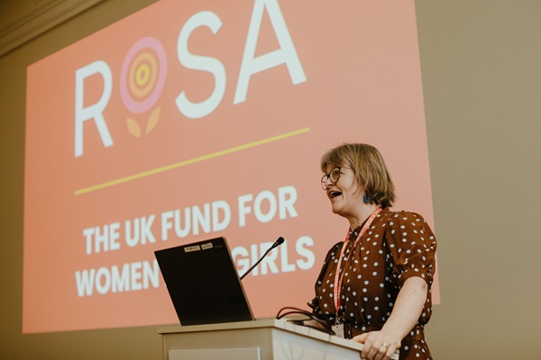 Woman speaking at a podium, with 'Rosa, the UK fund for women and girls' projected on the wall behind her.