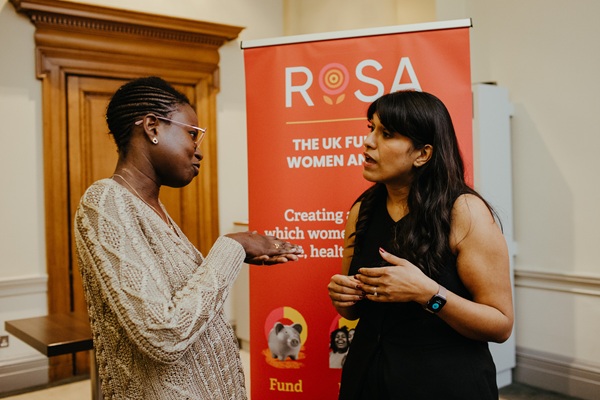 Two women in conversation in front of a red Rosa banner.
